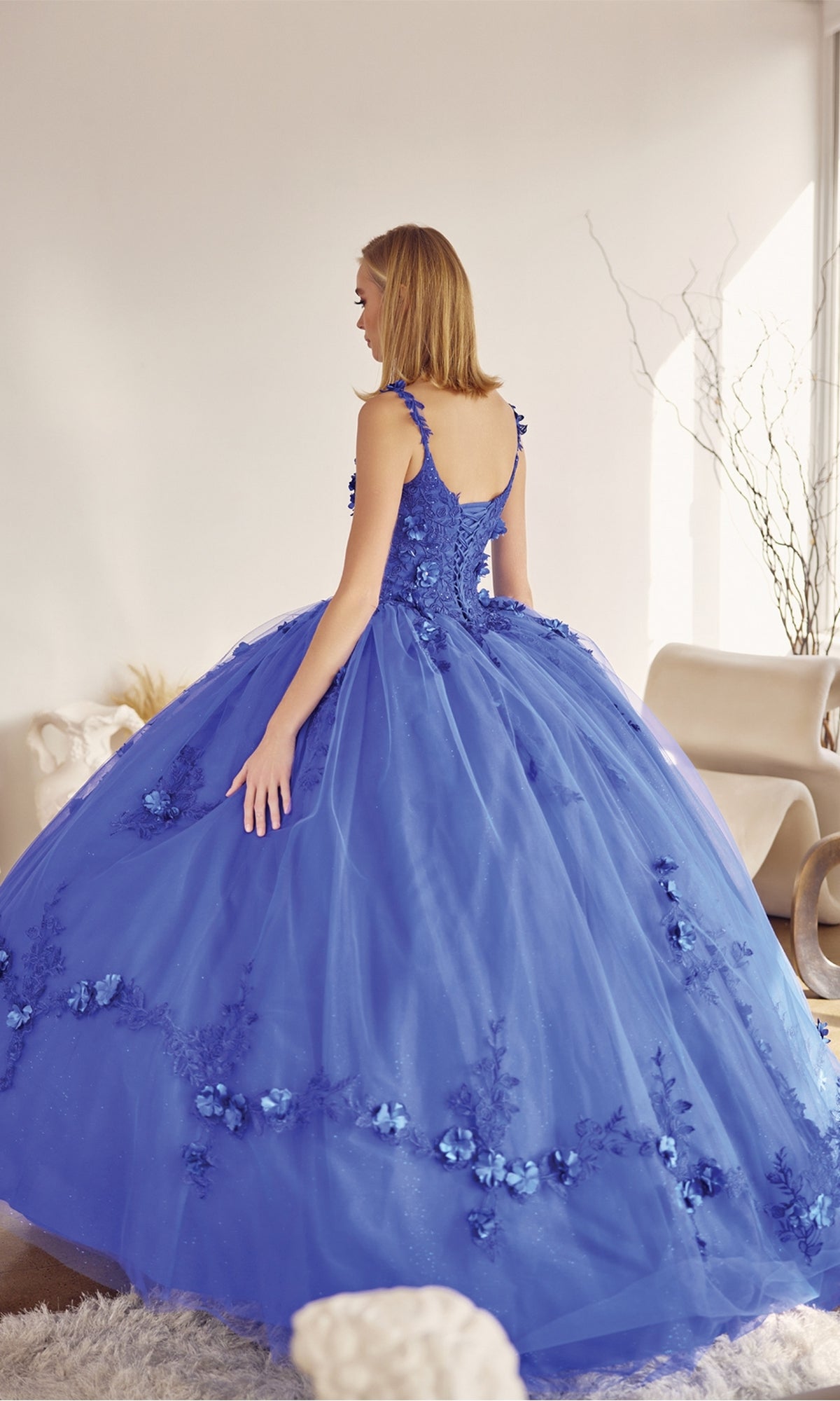 A woman in the Juliet 1436 Glitter Quinceañera Dress with Cape, featuring a voluminous blue skirt and floral appliqués, stands indoors facing away from the camera.